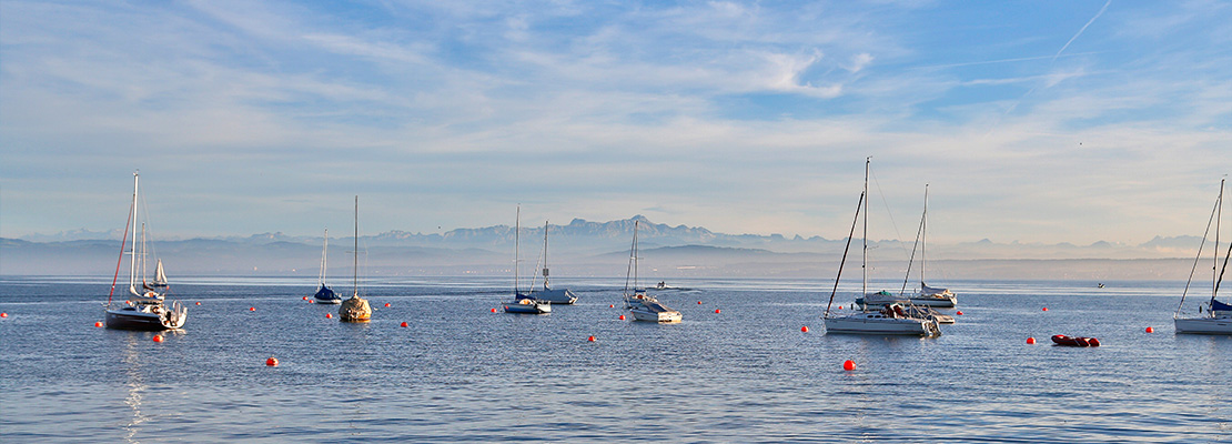 Panoramabild über den Bodensee mit Segelschiffen, im Hintergrund erkennt man einen Gebirgszug