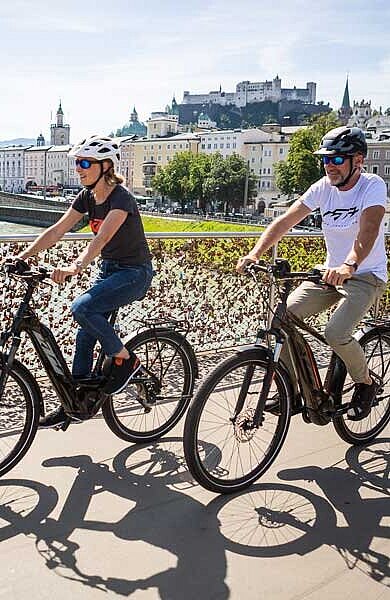 Mann und Frau auf E-Bikes fahren über die Salzachbrücke in Salzburg, dahinter die Festung