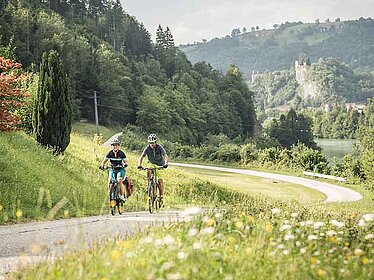 2 cyclists on a lonely street surrounded by meadows. In the back mountains and a castle