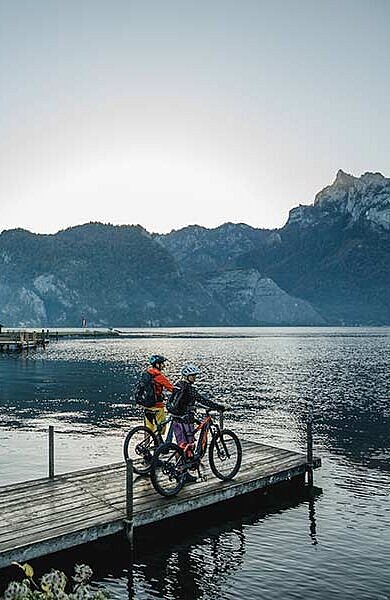 2 Personen mit Bikes am Steg am See, im Hintergrund Traunkirchen