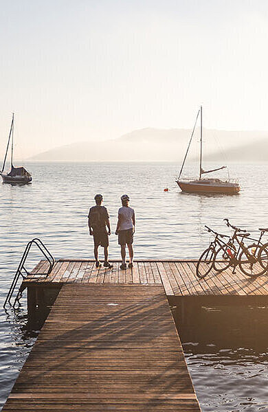 2 cyclists on a stage. boats on the lake, mountains in the far.