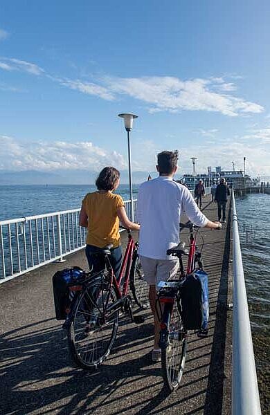 Zwei Radfahrer stehen auf einem Steg und blicken auf den Bodensee