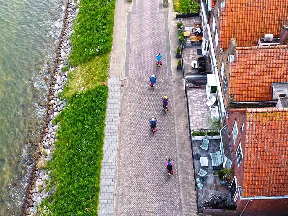 5 Radfahrer am Radweg, links das Meer, rechts eine Häusezeile aus der Vogelperspektive
