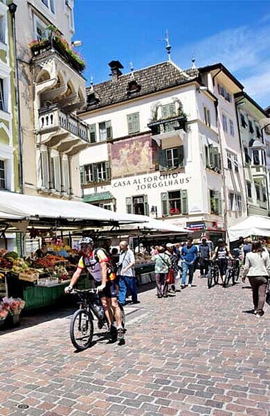 2 Radfahrer am Marktplatz von Bozen, vor Marktständen, im Hintergrund die Häuser der Altstadt