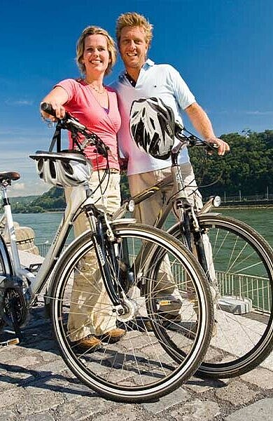 Ein Paar steht bei ihren Fahrrädern, den Helm am Lenker befestigt. Im Hintergrund die Mosel.