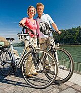 Ein Paar steht bei ihren Fahrrädern, den Helm am Lenker befestigt. Im Hintergrund die Mosel.