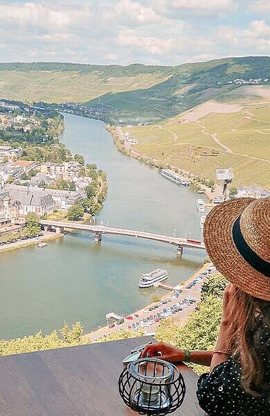Eine Dame mit Hut sitzt am Panoramafenster auf der Burg Landshut und blickt über die Stadt Bernkastel-Kues und die Mosel.