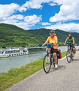 3 Radfahrer fahren am Radweg, im Hintergrund die MS Vivienne auf der Donau