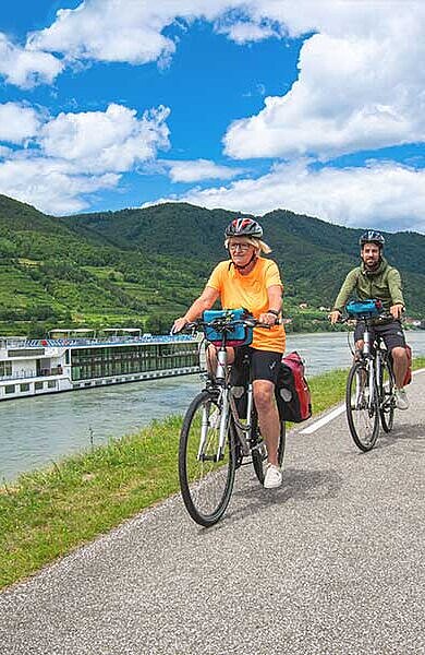 3 Radfahrer fahren am Radweg, im Hintergrund die MS Vivienne auf der Donau