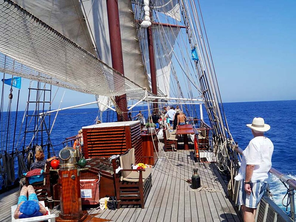 Oberdeck mit gesetzten Segeln, einer Dame am Liegestuhl und einem Mann, der aufs Meer blickt