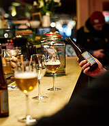 Biergläser und -flaschen stehen auf einer Theke, ein Mann hält eine Flasche in der Hand und liest das Etikett