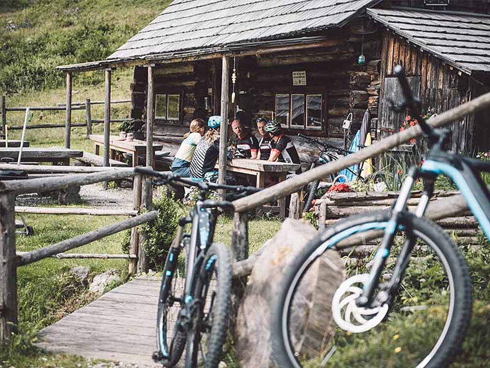 eine Hütte, davor Tische mit Bikern, im Vordergrund Bikes an ein Geländer gelehnt
