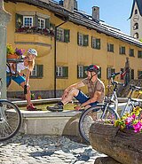 zwei Radfahrer machen Pause in der Sonne am Brunnen im Dorfplatz in Zuoz