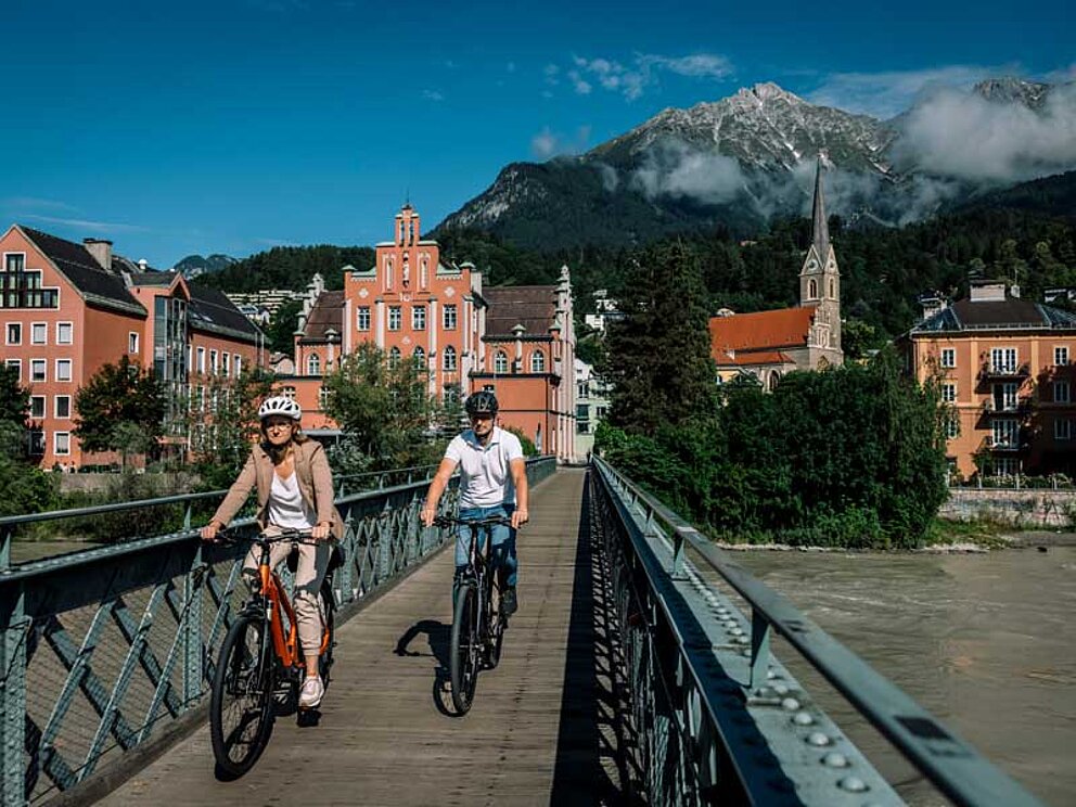 Mann und Frau auf Fahrrädern, die in Innsbruck über eine Brücke radeln. im Hintergrund die Alpen.